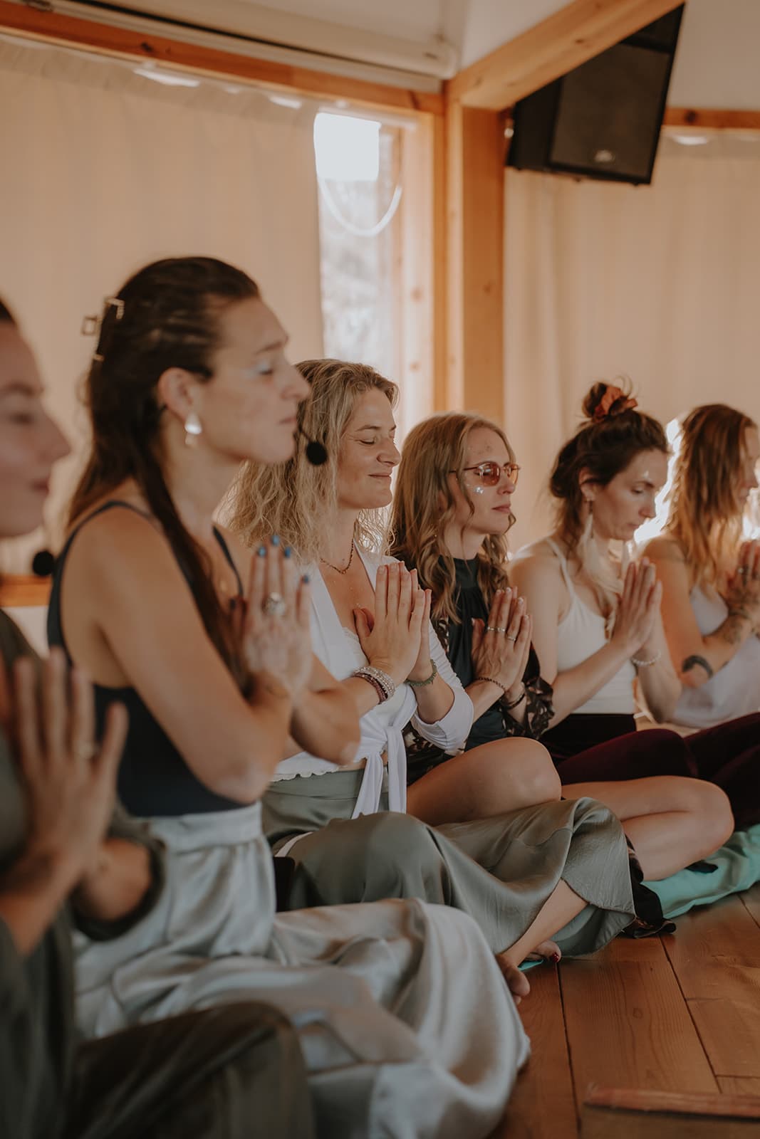 A row of women sitting cross legged on a wooden floor, eyes closed and palms together, during a ceremony
