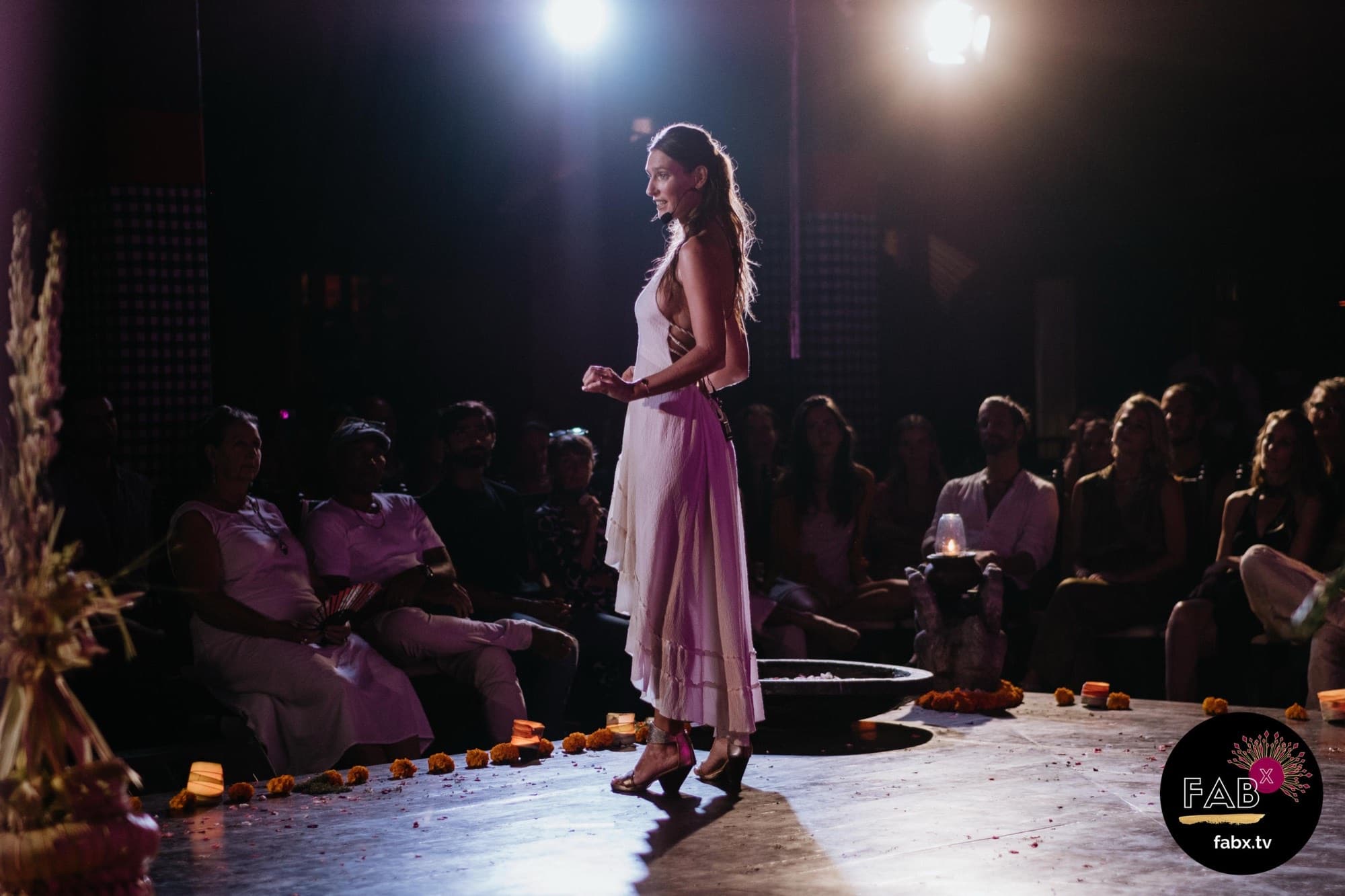 Arja Hendrikx in a white dress speaking under a spotlight to a seated audience, marigolds along the edge of the stage