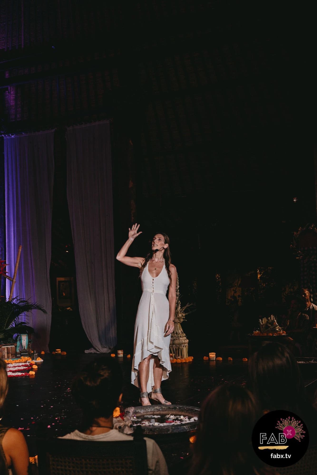 Arja Hendrikx in a white dress on a dark stage, one hand raised, audience in the foreground